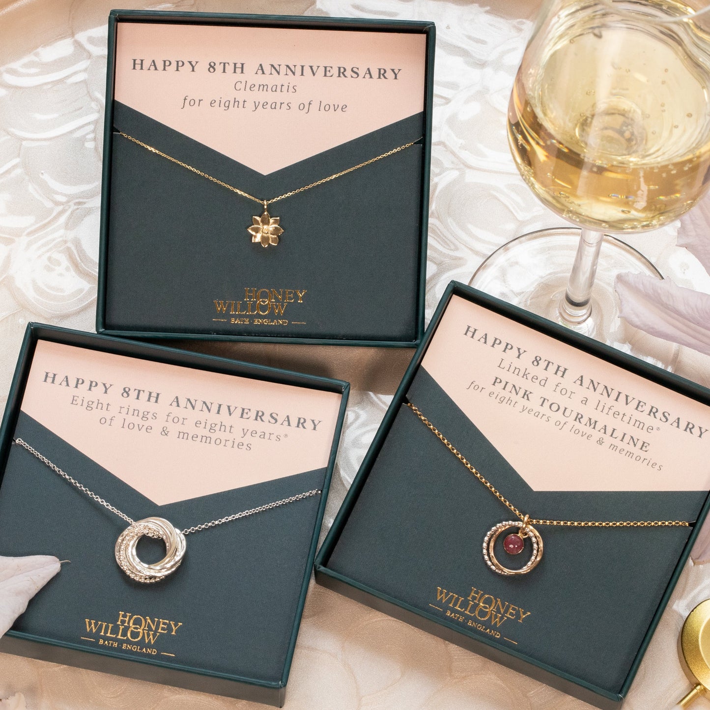 Three 'Happy 8th Anniversary' necklaces in boxes on a table with wine glasses and flowers.