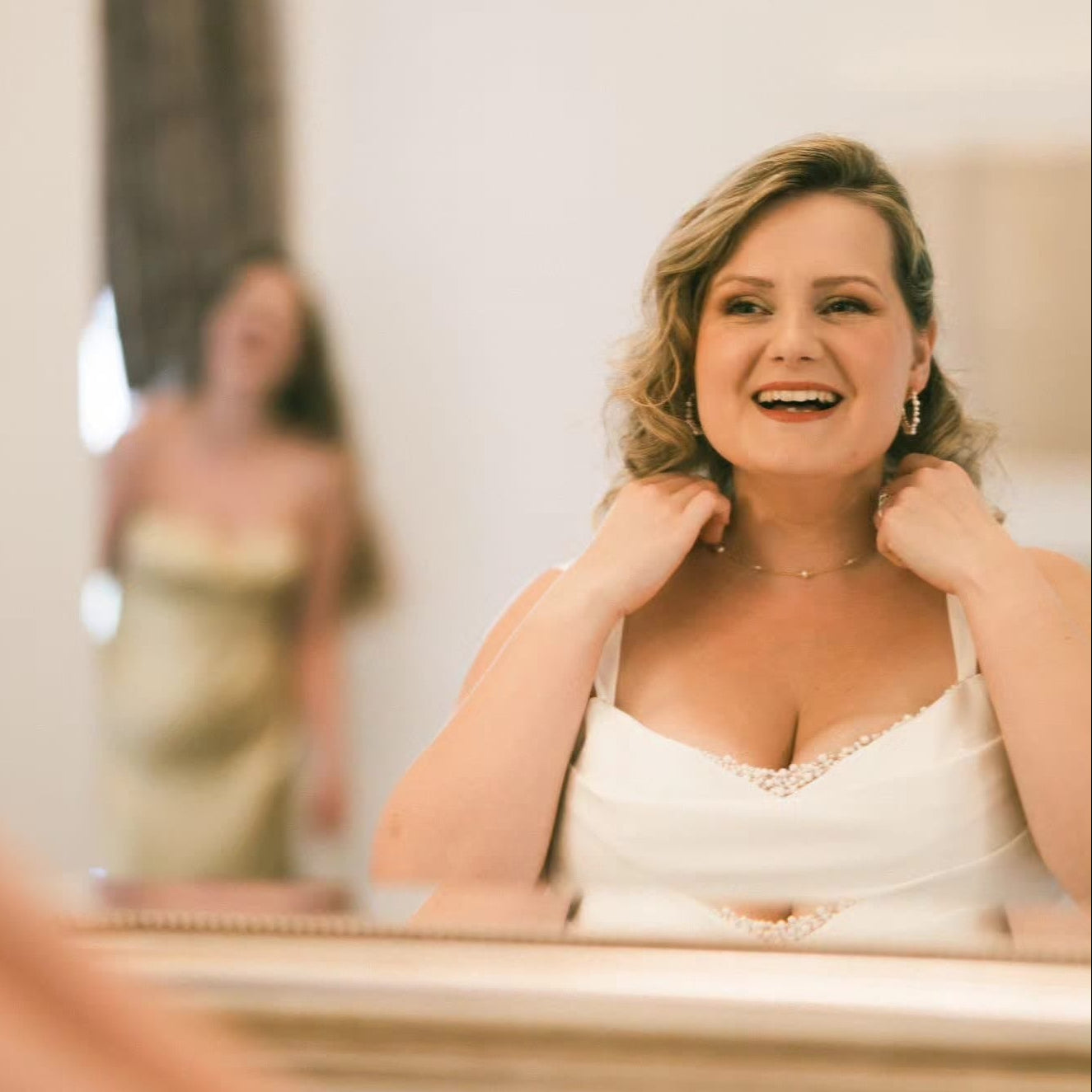 Woman in a white dress smiling in front of a mirror with another person in a gold dress in the reflection.